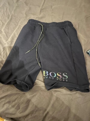 Svarta shorts från Hugo Boss - Säljer ett par svarta shorts från Hugo Boss med snyggt regnbågsskiftande logga på benet. De har elastisk midja med snörning och är gjorda i mjukt bomullsmaterial. Perfekta för chill eller streetwear, riktigt clean look.