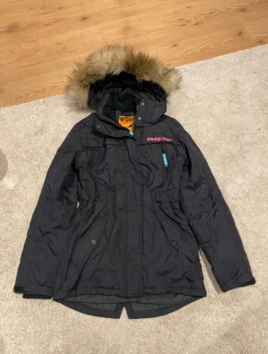 Svart skidjacka med fuskpäls från KappAhl - Svart skidjacka från KappAhl, med huva och fuskpäls. Jackan har dragkedja, knappar och flera fickor. Snygg broderad logga i rosa på bröstet och blå detalj vid fickan. Perfekt för kalla dagar och har en längre passform.