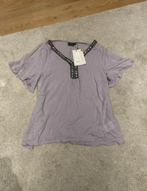 Grå blus med dekorerad v-ringning ZAY - Supersnygg grå blus från ZAY med korta ärmar och v-ringning. Halsringningen är dekorerad med svarta och silvriga pärldetaljer som ger en unik look. Blusen har lätt struktur i tyget och är perfekt för dig som vill sticka ut lite extra.