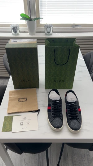 Gucci GG Supreme sneakers svart/röd - Gucci sneakers i svart GG Supreme canvas med klassiska röda och marinblå ränder på sidorna. Skorna har vita gummisulor, svarta skosnören och röd metallic-detalj på hälen. Snyggt mönstrade med diskret GG-logga över hela skon. Kommer med originalkartong och dustbag.