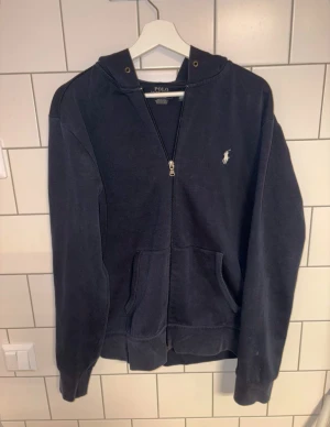 Polo Ralph Lauren zip - Snygg mörkblå hoodie från Polo Ralph Lauren med klassisk logga broderad i vitt på bröstet. Tröjan har huva, dragkedja framtill och två fickor. Tillverkad i mjuk bomull, perfekt för chill dagar. Passformen är normal och ärmarna är långa.