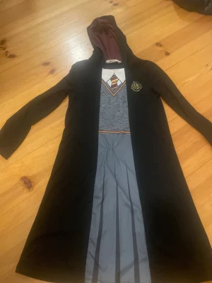 Hogwarts-inspirerad långklänning H&M - Säljer en Hogwarts-inspirerad långklänning från H&M med huva och långa ärmar. Klänningen har svart ytterdel, grå plisserad kjol och tryck med skjorta, slips och emblem på bröstet. Perfekt för dig som älskar Harry Potter och vill sticka ut med magisk stil.