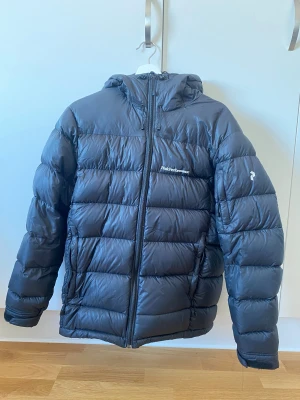 Svart Peak Performance dunjacka S - Svart dunjacka från Peak Performance i storlek S. Jackan har huva, quiltad design och dragkedja framtill. Perfekt för kalla dagar med sin fluffiga och varma känsla. Diskret logga på bröstet och ärmen. Snygg och enkel att matcha med allt.
