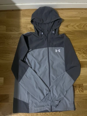 Grå vindjacka från Under Armour - Snygg grå vindjacka från Under Armour med huva och hel dragkedja. Jackan har två olika grå nyanser, logga på bröstet och stor Under Armour-tryck på ryggen. Perfekt för träning utomhus, med långärmade ärmar och lätt syntetmaterial. Kvitto finns.