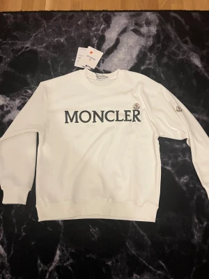 Vit sweatshirt från Moncler S - Vit sweatshirt från Moncler med stort svart logotryck på bröstet och broderad Moncler-patch på ärmen. Klassisk rund hals och ribbade muddar vid ärmslut och nederkant. Perfekt för dig som gillar stilrena och exklusiva plagg. 