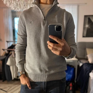 Grå half zip tröja Polo Ralph Lauren - Snygg grå långärmad tröja från Polo Ralph Lauren i mjuk pima bomull. Klassisk half zip med hög krage och broderad logga på bröstet. Perfekt till jeans för en clean och stilren look.