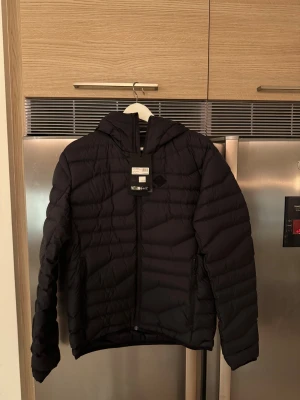 Svart dunjacka från J.Lindeberg - Svart dunjacka från J.Lindeberg med huva och snygga quiltade detaljer. Jackan har dragkedja framtill och logotyp på huvan. Perfekt för kalla dagar och har en modern, clean look som funkar till streetwear. Modellen heter Cliff Light Down Hood.