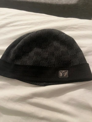 Louis Vuitton mössa  - Snygg svart mössa från Louis Vuitton med diskret rutigt mönster och LV-logga framtill. Tillverkad i mjuk ullmix med bred ribbad kant för extra komfort. Perfekt accessoar för att hålla stilen under kyliga dagar.