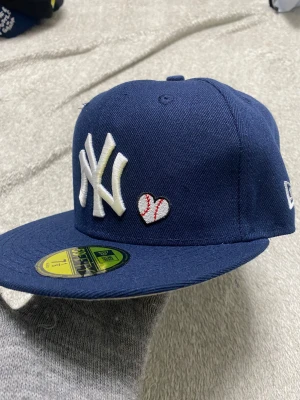 New Era Yankees 59FIFTY keps marinblå - Säljer en marinblå New Era 59FIFTY keps med New York Yankees-logga framtill, broderat hjärta i basebollstil och patch på sidan. Kepsen har platt skärm, broderad text bak och klassisk Cooperstown Collection-märkning. Perfekt för dig som gillar sportig streetstyle.