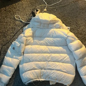 Vit Moncler pufferjacka med huva - Säljer en snygg vit pufferjacka från Moncler med ikonisk logga på ärmen. Jackan har huva, dragkedja framtill och praktisk ficka med knapp. Materialet är glansigt och jackan är dunfylld för extra värme. Perfekt för kalla dagar och riktigt clean stil. Tagg funkar 