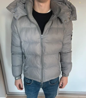 Grå pufferjacka från NVLTY med huva - Säljer en grå pufferjacka från NVLTY i storlek S. Jackan har en avtagbar huva.