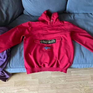 Röd hoodie från Napapijri med huva - Säljer en röd hoodie från Napapijri med stor huva och lång ärm. Tröjan har en cool ficka framtill med logotyp och norsk flagga. Ribbad nederkant och ärmslut ger en schysst passform. Perfekt för dig som gillar streetwear och snygga detaljer.