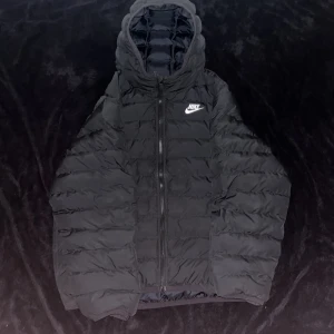 Nike jacka - Svart pufferjacka från Nike i storlek XL (158-170 cm). Jackan har huva, dragkedja framtill och klassisk Nike-logga på bröstet. Tillverkad i syntetmaterial och har en quiltad design som håller värmen under kalla dagar. Kom med prisförslag. Jackan passar både till ungdomar 13-15 år och även storlek S.