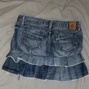 Jeanskjol med volanger - Säljer en cool jeanskjol med volangdetaljer och råa kanter. Kjolen har klassiska jeansfickor både fram och bak samt tre knappar framtill. Perfekt för dig som gillar en edgy och trendig stil.