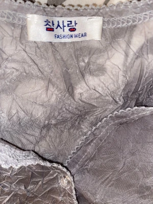 Grå transparent blus från Fashion Wear - Säljer en grå transparent blus med crinkle-effekt från Fashion Wear. Blusen har v-ringning, trekvartsärm och knappar framtill. Materialet är tunt och skimrande, vilket ger en cool och edgy vibe. Perfekt att styla med jeans eller kjol för en trendig look.