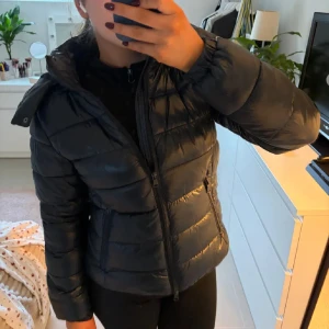 Svart Vinterjacka  - Snygg svart pufferjacka med glansig finish och avtagbar huva. Pris kan diskuteras 