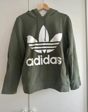 Grön Adidas hoodie med logga - Säljer en grön hoodie från Adidas med det klassiska trefoil-loggan i vitt framtill. Tröjan har huva med snörning och långa ärmar. Perfekt för både vardag och chill dagar