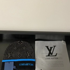  svart mössa med blå kant - Svart och grå mössa med klassiskt monogrammönster och broderad blå logga framtill. Mössan har en blå kant och är stickad i mjukt material som känns lyxigt och skönt. Perfekt för dig som vill ha en exklusiv look även när det är kallt.