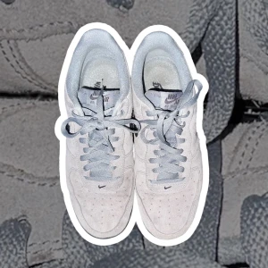 Nike Air Force 1 grå mocka sneakers - Snygga Nike Air Force 1 sneakers i ljusgrå mocka med matchande grå snörning och svart swoosh på sidan. Klassisk rund tå och perforerade detaljer på ovansidan för extra stil. Platt svart sula ger en clean look som funkar till allt.