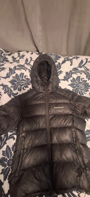 Svart Peak Performance dunjacka - Svart dunjacka från Peak Performance med huva och dragkedja framtill. Jackan har en quiltad design med breda paneler och snörning vid huvan. Perfekt för kalla dagar och har en clean look som funkar till det mesta.