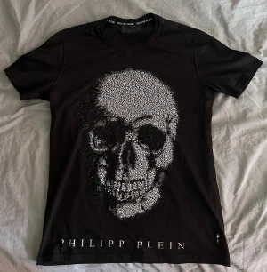 Philipp Plein - Säljer en svart t-shirt från Philipp Plein med en stor dödskalle framtill i rhinestone mönster och logga både fram och bak. T-shirten har rund hals och korta armar.