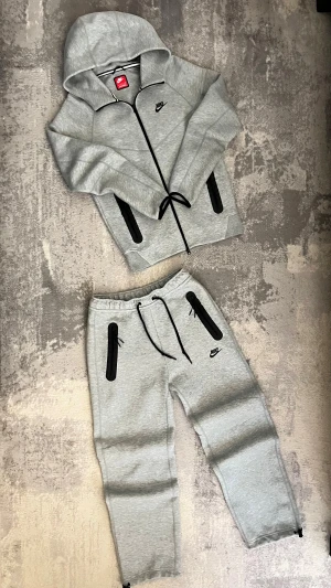 Grå mjukisbyxor och tröja från Nike  - Snygga grå mjukisbyxor och tröja från Nike tech med svarta dragkedjor på fickorna och justerbar midja med snörning. Byxorna har en avslappnad passform och är perfekta för chill eller träning. Materialet är mjukt och skönt.