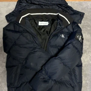 Mörkblå pufferjacka från Calvin Klein - Säljer en mörkblå Calvin Klein pufferjacka i väldigt fint skick. Jackan har vit CK-logga på bröstet och ärmen samt en avtagbar huva som enkelt kan tas av med dragkedja. På insidan av kragen finns en vit detalj som ger jackan en snygg och lite mer unik känsla.  Den är varm, bekväm och passar perfekt under höst- och vintermånaderna. En stilren jacka som fungerar till det mesta.  Hör gärna av dig vid minsta lilla funderingar!😊