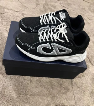 svarta sneakers - Säljer ett par sneakers i svart med silvriga detaljer och vit sula. Skorna har en sportig vibe med tydlig logga på sidan, vita snören och meshpartier för extra komfort. Perfekta för dig som vill sticka ut med lyxiga och trendiga sneakers.