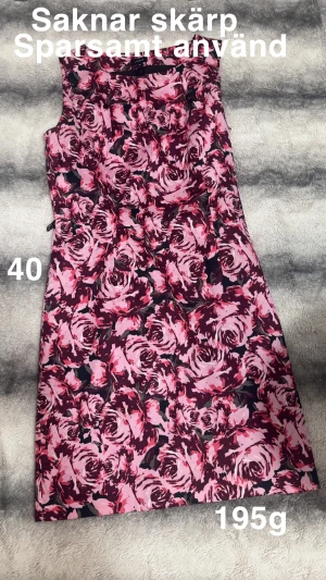 Blommig rosa klänning med bälte - Snygg ärmlös klänning från Lindex med rosa och vinröda rosor över hela plagget. Medföljer matchande bälte med rosett. Klänningen har en rak, kort siluett och dragkedja i sidan. Perfekt för dig som gillar färgstarka mönster och vill sticka ut.🌸 Sparsamt använd. Skärpet som hör till syns på andra bilden . Storlek 40