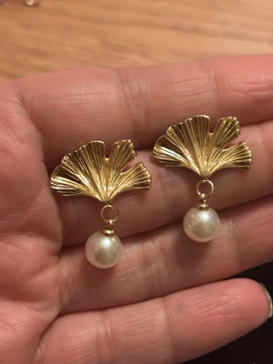 Stål Örhängen Ginko Pearl - Säljer ett par snygga örhängen stål metall med ett detaljerat gynko hänge och en rund vit pärla som dinglar under. Perfekt för dig som gillar unika smycken med naturinspirerad design och elegant touch.