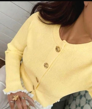 Gul stickad tröja  med guld knappe  - Snygg gul stickad tröja med långa ärmar och vita volangdetaljer vid ärmslut och nederkant. Koftan har guldfärgade knappar framtill men dom går inte att knäpp upp utan är bara en design , och en rund halsringning. Perfekt för dig som vill ha en söt och fräsch look 