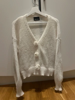 Vit kofta med blomknappar från Pieces - Superfin vit kofta från Pieces med stickad struktur och tre unika blomformade knappar framtill. Modellen har v-ringad hals, ribbade muddar och är långärmad. Perfekt för dig som gillar söta detaljer och vill ha en mysig look.