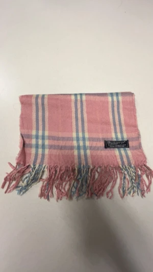 Rutig rosa halsduk från Burberrys - Säljer en klassisk rutig halsduk från Burberrys i mjuk ull. Färgen är ljusrosa med inslag av blått och beige, och den har fransar i ändarna. Perfekt accessoar för att lyfta din outfit med lite färg och stil.