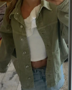 Olivgrön jeansjacka med stora fickor - Säljer en olivgrön jeansjacka med stora framfickor och klassisk krage. Jackan har knäppning framtill med metallknappar och raka, långa ärmar. Perfekt för lager-på-lager och enkel att styla till olika outfits. 