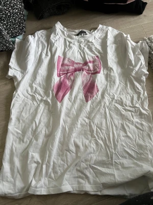 Vit t-shirt med rosa rosett new yorker - Säljer en vit t-shirt från New yorker med en stor rosa rosett framtill. T-shirten har korta ärmar och rund hals. Perfekt för dig som gillar söta detaljer och vill ha en enkel men ändå unik look.