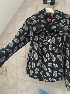 Svart leopardmönstrad kappa från Derhy - Snygg svart kappa från Derhy med grått leopardmönster och stora svarta knappar. Jackan har bred krage, lång ärm och är tillverkad i ett mjukt ullmaterial. Perfekt för dig som vill sticka ut med ett coolt mönster och trendig look.