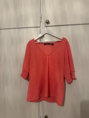 Korallrosa blus med v-ringning - Säljer en korallrosa blus från H&M med v-ringad hals och korta ärmar med smockdetalj. Blusen har en lätt skrynklig struktur i tyget och är luftig och bekväm. Perfekt för dig som gillar färg och vill ha en chill topp till sommaren.