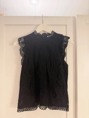 Svart spetsblus med hög krage - Supersnygg svart blus i spets med hög krage och ärmlös design. Blusen har detaljerade spetsmönster och vackra kanter längs axlar och nederkant. Dragkedja i ryggen för enkel på- och avtagning. Perfekt för dig som gillar en elegant och trendig look.
