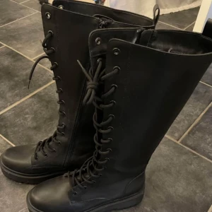 Svarta höga snörade boots - Säljer ett par svarta höga boots med snörning hela vägen upp och grov platt sula. Skorna är tillverkade i skinn och har en rund tå. Perfekta för dig som gillar en edgy stil och vill ha något som sticker ut. inte använda då dom inte passade mig