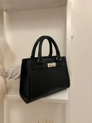 Svart handväska med silverdetaljer - Svart liten handväska i skinnimitation. Väskan har en avtagbar och justerbar axelrem och silverfärgade metalldetaljer 👜🤍