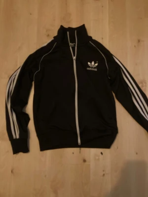 Svart Adidas track jacket med vita ränder - Svart Adidas track jacket med klassiska vita ränder längs ärmarna och vit dragkedja. Jackan har hög krage, lång ärm och Adidas logga på bröstet. Tillverkad i polyester för en sportig vibe. Perfekt för dig som gillar streetwear och sportig stil.