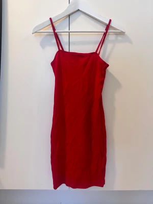 Röd kort klänning med smala axelband - Säljer en röd kort klänning med smala axelband och rak passform. Klänningen har en enkel och stilren design som ger en snygg siluett. Perfekt för dig som gillar minimalistisk stil och vill sticka ut med färg.