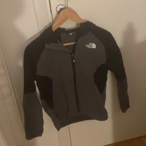 Svart och grå fleecejacka North Face - Säljer en svart och grå fleecejacka från The North Face med huva och hel dragkedja framtill. Jackan har långärmade ärmar och logga på bröstet. Perfekt för kyliga dagar och har en sportig look med kontrastpaneler.