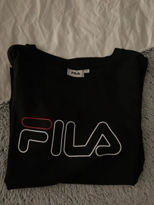 Svart FILA t-shirt med logga - Svart t-shirt från FILA med stor vit och röd logga framtill. Klassisk rund halsringning och korta ärmar. Skön passform och tillverkad i mjuk bomull, perfekt för en sportig och avslappnad stil. Aldrig använd men lapparna är avklippta💗