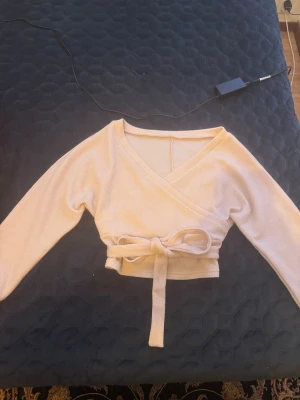 Beige omlottopp med knytband - Superfin croppad omlottopp i ljus beige stickad kvalitet. Toppen har långa ärmar, v-ringning och ett brett knytband i midjan som ger en snygg siluett. Perfekt för dig som gillar mjuka material och stilrena detaljer.