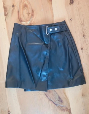 Svart omlottkjol i skinnimitation Divided - Snygg svart kort omlottkjol från Divided i skinnimitation. Kjolen har en cool detalj med bälte och silverfärgat spänne i midjan. Perfekt för dig som gillar en edgy stil och vill sticka ut lite extra.