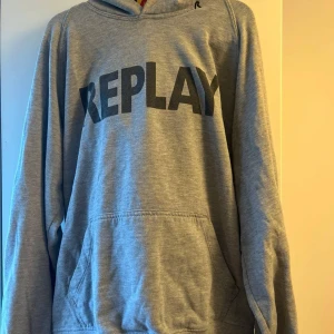 Grå hoodie från Replay med tryck - Snygg grå hoodie från Replay med stor svart logga framtill. Klassisk känguruficka och huva, perfekt för chill dagar. Mjuk bomullsblandning och långärmad modell som funkar året runt. Enkel och stilren design som passar till det mesta.