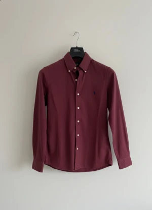  Ralph Lauren skjorta vinröd S - Vinröd skjorta från Ralph Lauren i klassisk passform, med knappar framtill och på kragen. Skjortan har lång ärm och ikonisk broderad logga på bröstet. Tillverkad i mjuk bomull som känns skön mot huden. Perfekt för dig som gillar stilrena och tidlösa plagg. Inga defekter och tröjan är i ny skick. Hör av dig vid frågor/prisförslag😇🙌