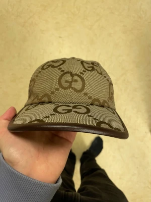 beige jumbo keps med monogram - Snygg beige jumbo keps med monogram över hela kepsen. Skärmen och justerbar rem bak är i brunt skinn, vilket ger en lyxig känsla. Perfekt för dig som vill sticka ut med en ikonisk accessoar från ett av världens mest kända modehus.