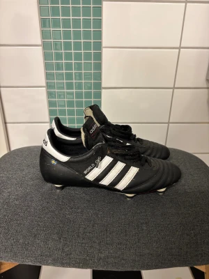 Adidas World Cup fotbollsskor svart/vit skruvdobb - Klassiska Adidas World Cup fotbollsskor i svart skinn med vita detaljer och de ikoniska tre ränderna på sidan. Skorna har snörning och metallskruvdobbar för extra grepp på planen. Snygg retrostil med rund tå och Adidas-logga på plösen och hälen. Aldrig använda ba på en träning. Skicka vid frågor. ( priset kan diskuteras)